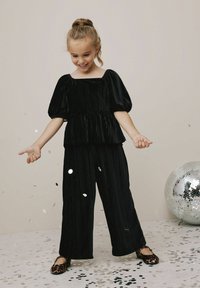 Czarna welurowa bluzka z bufkami i kwadratowym dekoltem w połączeniu z czarnymi plisowanymi spodniami; nosząca baleriny w panterkę. Konfetti rozsypane na ziemi.