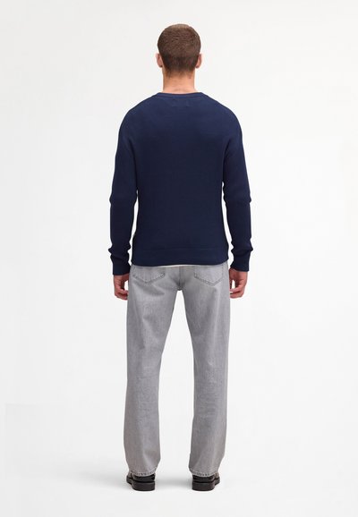 Mand stående med ryggen til iført en marineblå ribstrikket sweater, lysegrå jeans med lige ben og sorte sko på en hvid baggrund.