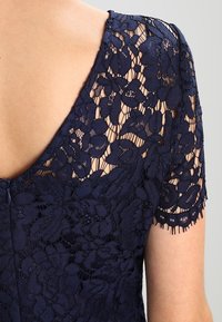 Abito in pizzo floreale blu navy con maniche corte e dettaglio a V sulla schiena che mostra la pelle attraverso il tessuto trasparente.