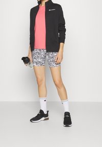 Svart zip-up jacka, rosa t-shirt, cykelshorts med svart och vit mönstring, vita strumpor med gröna ränder, svarta träningsskor, håller en vattenflaska.