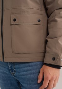 Chaqueta impermeable beige con cremallera en la parte delantera, dos grandes bolsillos en el pecho con botones negros y tela texturizada. Combinada con jeans azules.