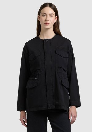 Mujer con una chaqueta negra abotonada con cuatro bolsillos delanteros con solapa, frente a un fondo gris liso.