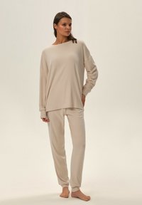 Beżowy zestaw odzieży domowej składający się z długiego topu z szerokim dekoltem i luźnym krojem, w komplecie z dopasowanymi spodniami o tapered kroju. Gładka tekstura.