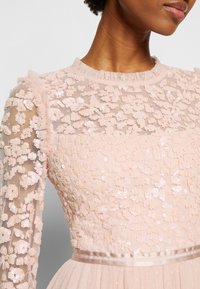 Gros plan d'une personne portant une robe légère rose clair transparente avec des ornements floraux en sequins et un col et des manches à volants.