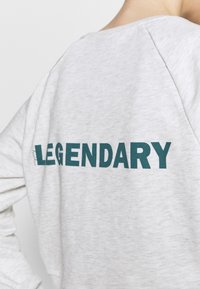 Světlě šedá mikina s raglánovými rukávy, na zádech vytištěné slovo "LEGENDARY" v tyrkysové barvě. Hladká, měkká textura látky.