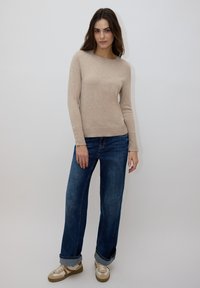 Pull en maille beige avec col rond et manches longues, associé à un jean bleu à jambes larges. Les chaussures présentent un design beige clair et blanc avec des lacets.