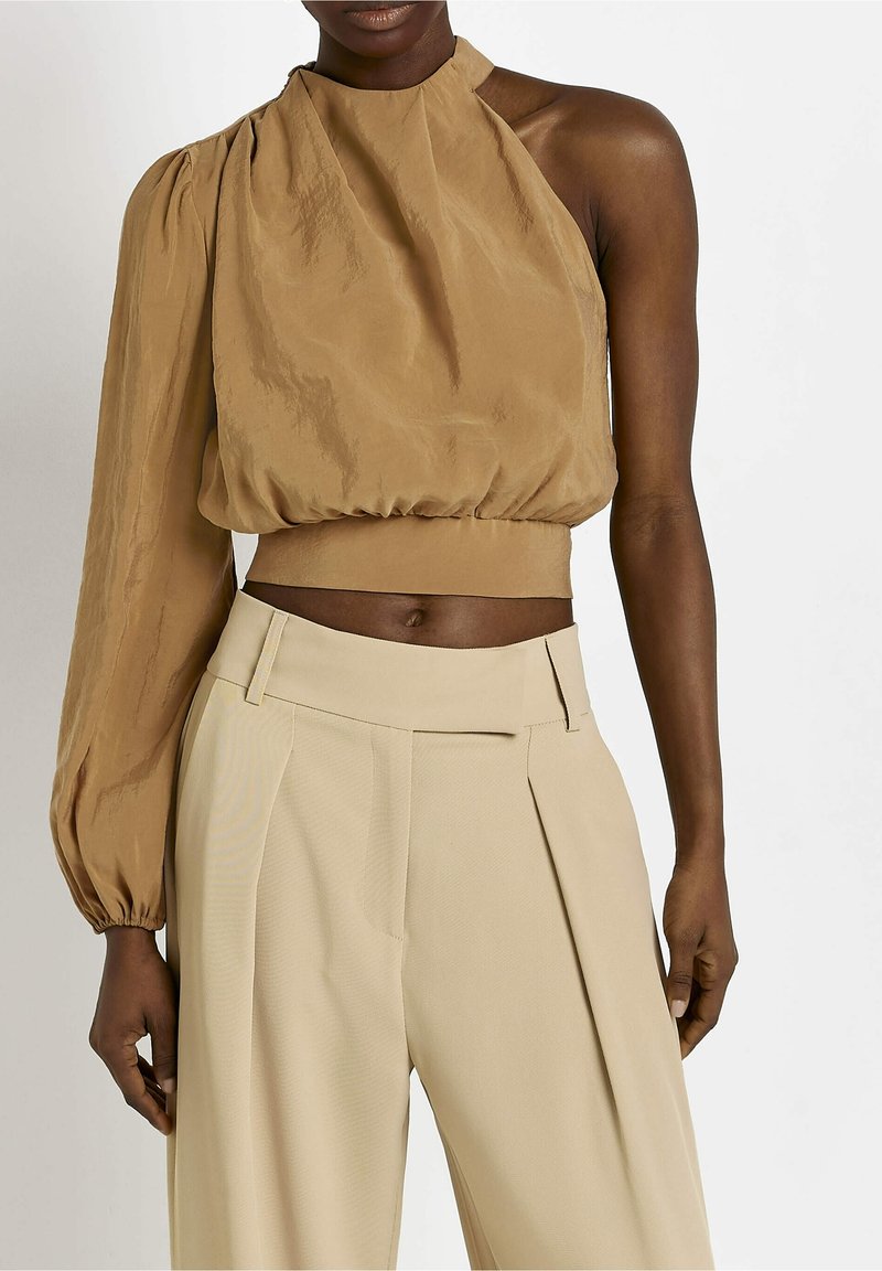 Brun enaxladad blus med en strukturerad yta, voluminös puffärmar och en figursydd midja. I kombination med beige höga, veckade byxor.