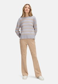 Frau, die einen grauen Pullover mit beigen Streifen, beige Hosen und weiße Sneaker vor einem weißen Hintergrund trägt.