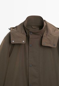 Brązowa wodoodporna kurtka z wysokim kołnierzem, zatrzaskami i odpinanym kapturem. Tkanina ma gładką, matową fakturę oraz czyste linie.