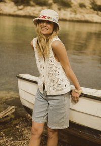 Gehaakte crèmekleurige vest met een sluiting aan de achterkant met knopen, gedragen met lichte denim shorts. Het model draagt een patronen bucket hat en een armband.