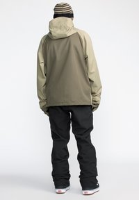 Ski jacket in beige and black, featuring a hood and long sleeves. Paired with black pants and black footwear, showing a relaxed fit.