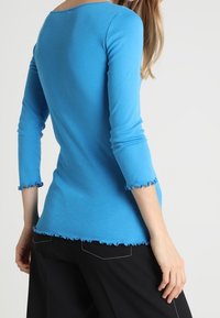 Långärmad, figurnära topp i ljusblå bomull med båthals och vågiga kanter vid ärmarna och nederkanten. Matchad med svarta byxor.