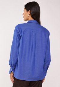 Camicia blu con bottoni, maniche lunghe, pieghe sul retro e polsini con bottoni. Tessuto liscio con colore uniforme. Dettagli hardware minimi.