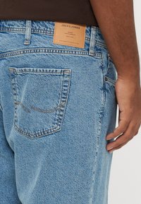 Gros plan d'une personne portant un jean Jack & Jones bleu clair, montrant la poche arrière, un patch en cuir de la marque et la main droite reposant sur le côté.