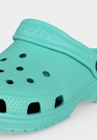 Gros plan d'un sabot Crocs turquoise montrant des trous de ventilation, une surface texturée et une bride de talon réglable.