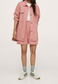 Różowa oversize'owa kurtka i dopasowane szorty z miękkiego materiału, z czterema przednimi kieszeniami i detalami z guzikami. Do tego szare skarpetki i trampki.