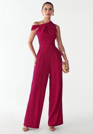 Bordeauxrød heldragt med off-shoulder design og knudedetalje. Flydende vidbenet silhuet, lavet af letvægtsstof, suppleret med guldtilbehør.