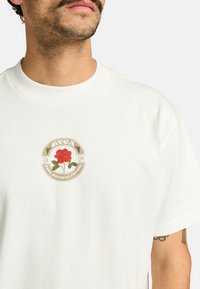 Camiseta blanca de algodón con un logo redondo que presenta una rosa roja y hojas verdes, acentuado por el texto "RVCA" y "PASADO.PRESENTE.FUTURO."