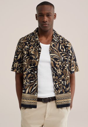 Man draagt een zwart en beige kortebroek overhemd met bloemenpatroon over een wit T-shirt, met beige broek en zwarte riem.