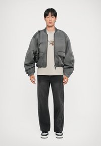 Gray bomber jacket with voluminous sleeves, worn over a beige graphic t-shirt and black jeans with side stitching, paired with black and white sneakers.