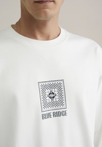 Balta palaidinė su kvadratine gėlių raštu tamsiai mėlyna spalva ir tekstu "BLUE RIDGE" po dizainu. Minkšta, lygi audinio tekstūra.