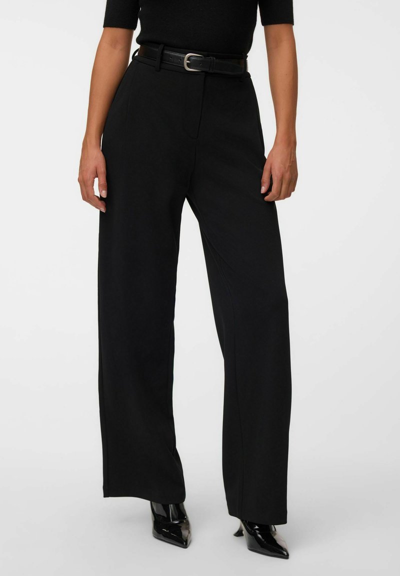 Femme portant un pantalon noir taille haute à jambes larges avec une ceinture noire, associé à des talons noirs brillants à bout pointu et un haut noir.