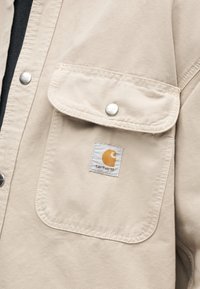 Μπεζ τσέπη σε μπουφάν Carhartt με καπάκι και κούμπωμα με τρουκ, με μικρό λευκό και πορτοκαλί λογότυπο Carhartt.