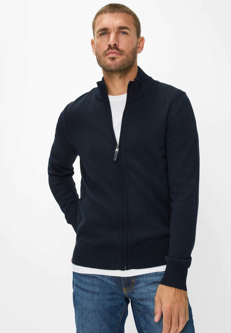 Marineblauer Reißverschluss-Pullover mit geripptem Kragen und Bündchen, aus weichem Strickmaterial gefertigt. Trägt man über einem weißen T-Shirt und kombiniert mit blauen Jeans.