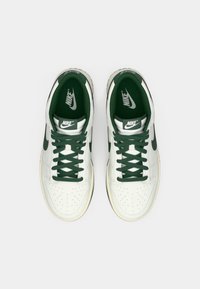 Pár bielych a zelených Nike tenísk s tmavozelenými šnúrkami, logo Nike na jazyku a vnútornom sole, zobrazené zhora na bielom pozadí.