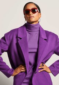 Lila Wolljacke mit Reverskragen über einem passenden gerippten Rollkragenpullover. Dazu rote Cat-Eye-Sonnenbrille und Creolen.