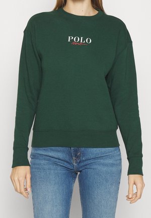 Femme portant un sweat-shirt vert foncé avec le logo « POLO Ralph Lauren », associé à un jean bleu, debout devant un fond clair uni.