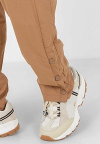 Bruine broeken met decoratieve knopen aan de manchetten, gecombineerd met beige en witte sneakers met donkere strepen en gestructureerde zolen.