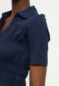 Άτομο με σκουρόχρωμο δέρμα που φοράει φόρεμα σε ναυτικό μπλε χρώμα με κοντά μανίκια, λεπτομέρειες με πιέτες υφάσματος και γιακά διπλωμένο, πάνω σε απλό φόντο.