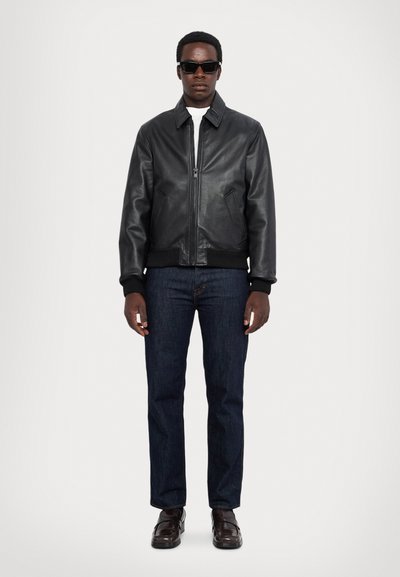 Chaqueta bomber de cuero negro con cremallera frontal, puños y dobladillo acanalados, combinada con jeans de mezclilla oscura y zapatos marrones. Modelo con gafas de sol.