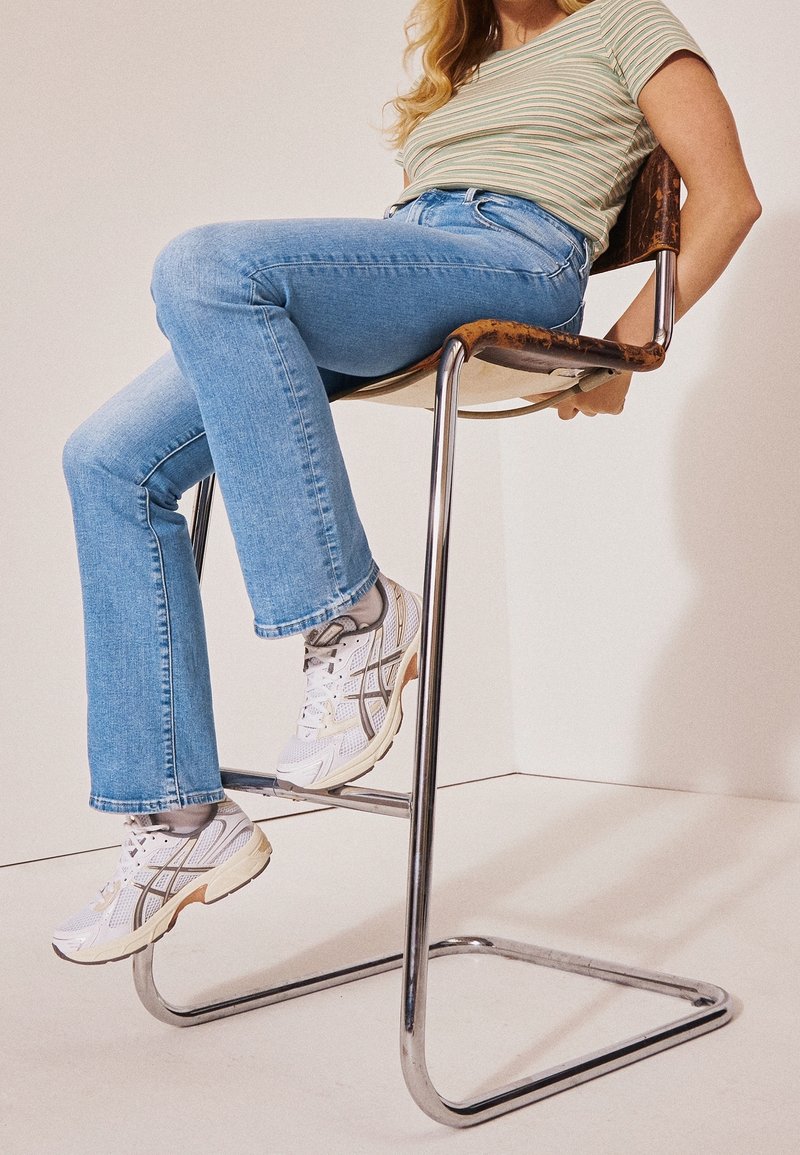 Femme portant une chemise rayée, un jean bleu et des baskets blanches, assise sur un tabouret de bar en métal avec un siège en cuir rouillé.