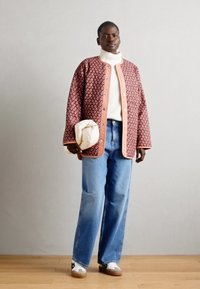 Vadderad jacka med röd och rosa mönstring, knappar, vit polotröja, ljusblå vida jeans och vita sneakers med bruna sulor.