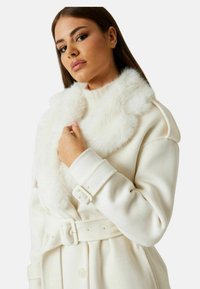 Cappotto in lana bianca con collo in pelliccia sintetica, vita con cintura e spalle strutturate. Tessuto morbido con chiusura frontale con bottoni e maniche arricciate.