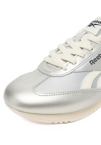Strieborná športová obuv s hladkým, lesklým povrchom, vybavená bielymi šnúrkami a výraznými detailmi. Dizajn obsahuje klasické logo Reebok.