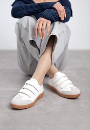 Sneakers bianchi con tre cinturini in velcro, punta rotonda e suola in gomma di colore marrone naturale, poggiati su un pavimento grigio.