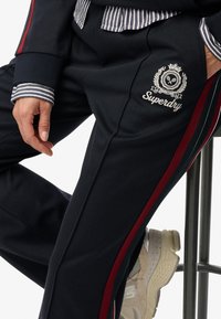 Granatowe spodnie dresowe z bordowymi paskami po bokach i haftowanym logo "Superdry", w połączeniu z koszulą w paski i jasnymi trampkami.