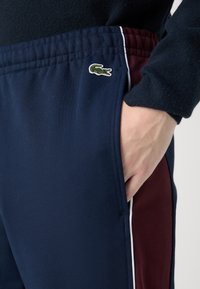 Jūras spēku sporta bikses ar bordo sānu paneļiem, elastīgu jostu un nelielu Lacoste krokodila logo priekšpusē. Gluda auduma tekstūra.