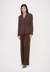 Hnedé na mieru šité sako s výrezmi na klopách a tromi gombíkmi, kombinované s voľne strihanými hnedými nohavicami a otvorenými hnedými lodičkami.