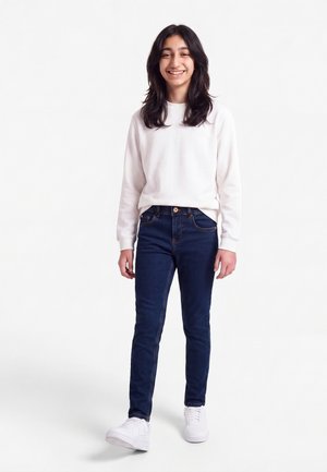 Χαμογελαστό άτομο με μακριά σκούρα μαλλιά, φοράει λευκή φούτερ μπλούζα, σκουρομπλε τζιν και λευκά αθλητικά παπούτσια, στέκεται μπροστά σε απλό λευκό φόντο.