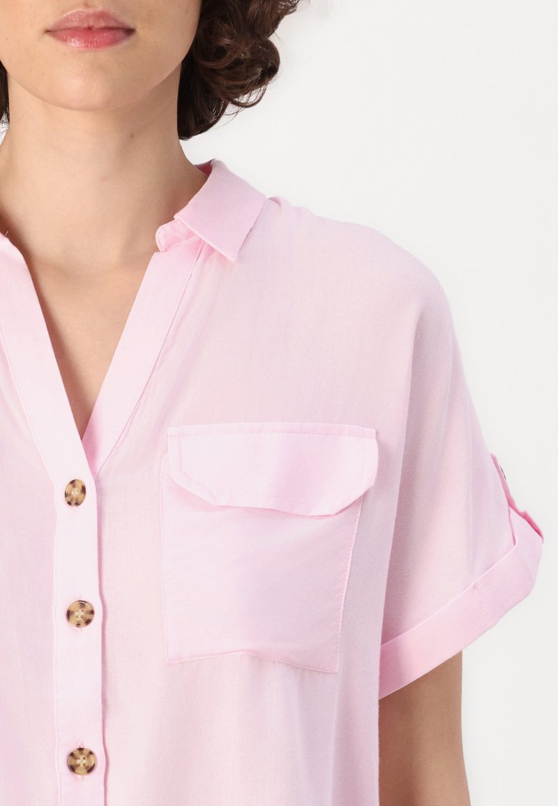 Blouse rose clair à manches courtes en tissu doux. Elle comporte un col, une poche avant et des détails de boutons le long de la patte de boutonnage.