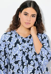 Kukallinen pusero tummansinisellä taustalla, jossa on vaaleansinisiä ja valkoisia abstrakteja kuvioita, pyöreä pääntie ja 3/4-pituiset hihat. Pehmeä kangastuntuma.