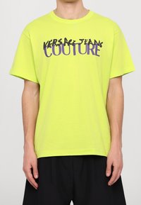 Neon žluté tričko vyrobené z bavlny, s odvážným grafickým designem s nápisem "VERSACE JEANS COUTURE" v purpurové a černé barvě. Krátké rukávy.