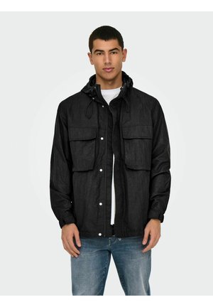 Veste noire légère avec capuche, deux poches avant, boutons-pression et un tissu légèrement texturé. Portée par-dessus un t-shirt blanc et un jean.
