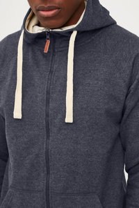 Mornarska zip-up dukserica s kremastožutim vezicama, koja se odlikuje mekanom teksturom, prednjim džepovima i malom bež oznakom blizu zatvaranja.