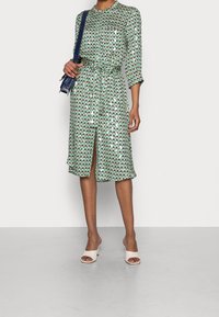 Vestito verde a fantasia con design triangolare, maniche lunghe, vita con lacci e uno spacco frontale. Abbinato a scarpe open-toe di colore chiaro.