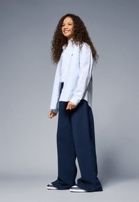 Camicia button-up di colore azzurro chiaro con colletto a punta, abbinata a pantaloni larghi di un navy scuro e sneaker bianche e nere. Capelli ricci.